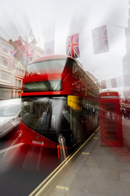 A red double-decker bus, displaying the route to Hammersmith, is parked on a street in London during daytime, with a traditional red telephone booth positioned next to the bus on the sidewalk. The scene includes decorative Union Jack flags hanging overhead, creating a festive atmosphere. Reflections of surrounding buildings and pedestrians can be seen on the bus's glossy exterior. In the background, there are historic commercial and residential buildings with ornate facades. The street appears busy, likely in a central area, with the bus and telephone booth indicating typical elements involved in urban house removals and local moving logistics. Hammersmith Man and Van’s presence is implied through the context of relocation services, with the scene showing an iconic part of city infrastructure used for transportation and packing during house moves. The lighting is natural, and objects like the bus, telephone booth, and flags are clearly visible, illustrating elements associated with urban home relocation and transport processes.