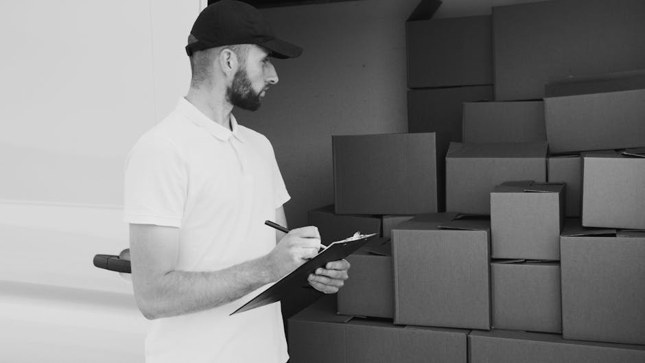A man in a white polo shirt and black cap is inside a storage area or a moving facility, standing in front of a large stack of uniformly shaped cardboard boxes. The boxes are neatly arranged against a dark wall, some stacked on top of each other on the floor, while others are placed on shelves or against the wall. The man is holding a clipboard and a pen, appearing to take inventory or check off items during a packing or loading process related to house removals or furniture transport. The environment is well-lit, with the focus on the man and the boxes, indicating a professional setting involved in packing, moving logistics, or home relocation activities, as typical for services provided by Hammersmith Man and Van.