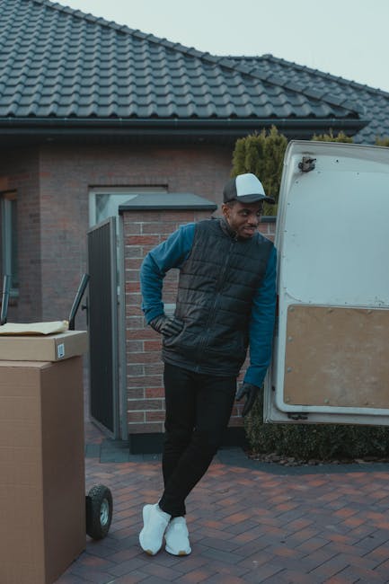 A man wearing a baseball cap, black gloves, a blue long-sleeve shirt, and a black padded vest leans against the side of a white moving van during a home relocation process. The van is parked on a bric