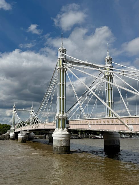 The image shows the Hammersmith Bridge spanning a river under a partly cloudy sky. The bridge features tall, ornate metal towers with spires, connected by a network of cables supporting the bridge deck. The span has a light pinkish color with decorative elements, and the base of the bridge is anchored on stone piers in the water. The water below is calm with minimal ripples, reflecting the structure above. Surrounding the bridge is a cityscape, with some buildings visible in the background. The scene captures the structural details of this historic suspension bridge, often involved in home relocation and furniture transport logistics, especially when navigating under or around such key infrastructure during house removals. Hammersmith Man and Van utilise such landmarks in their comprehensive removals and moving services to facilitate efficient access and planning for house moves near Hammersmith Bridge.
