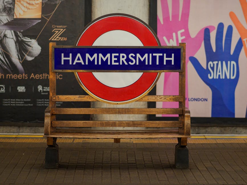 A wooden bench with a metal frame is positioned on a tiled pavement in Hammersmith, in front of a large circular red and white London Underground sign with a blue rectangular nameplate reading 'HAMMERSMITH.' Behind the sign, there are colorful posters; one features a black and white portrait with additional text, and another displays vibrant handprints with the words 'WE STAND' in white. The setting appears to be an indoor or covered area, possibly a station platform, during the day with even lighting. The scene may relate to home relocation or furniture transport, as Hammersmith Man and Van often provides removals and moving services, with the bench and signage serving as potential points for loading or temporary staging during a furniture move.