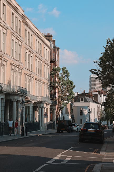 The image depicts a quiet residential street in Hammersmith W6 during daylight, featuring a row of multi-storey Victorian-style buildings with cream-colored facades, decorative molding, and wrought iron balconies. Several large cardboard boxes, some wrapped in plastic, are stacked on the sidewalk near the entrances, indicating packing for a home relocation. A wooden furniture piece and several smaller packed items are visible among the boxes. In the background, a white vehicle is parked, and further down the street, a black van with open rear doors is positioned near a loading area on the pavement, suggesting furniture transport and loading activities. Nearby, a few pedestrians walk along the sidewalk, and mature trees provide shade, contrasting the urban environment. This scene captures an active moving and packing process, consistent with professional removals services like those provided by Hammersmith Man and Van, specializing in home relocation and furniture transport, set against a typical London street environment with clear weather and natural lighting.
