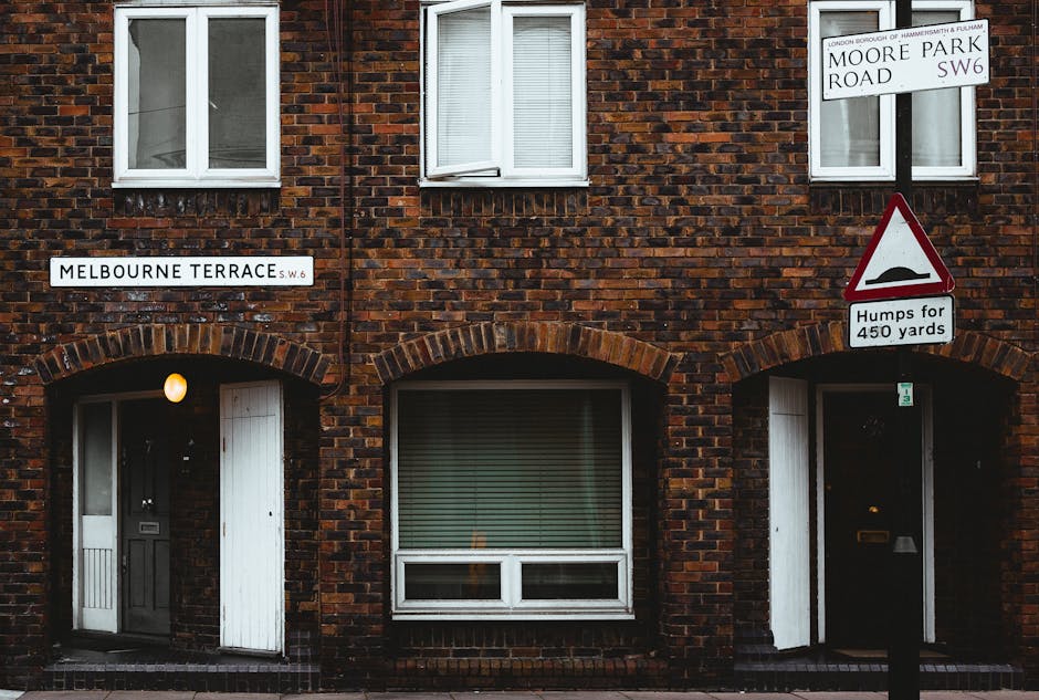 A brick residential building on Melbourne Terrace in Hammersmith W6, featuring two white-framed windows on the upper floor, with one window partially open, and two ground-floor entrance doors with small windows beside them. The left door is closed and painted white, while the right door is dark with a mailbox mounted nearby. Exterior lighting illuminates the entrance areas, and the building's brickwork is dark red with lighter mortar. A street sign indicates the location as Moore Park Road SW6, and another sign warns of Humps for 450 yards, with a red and white triangular traffic sign mounted on a black pole nearby. The scene is captured during daylight hours with even lighting, and the street signage and buildings identify the location as part of a walking route or residential area associated with house removals and relocations facilitated by Hammersmith Man and Van.