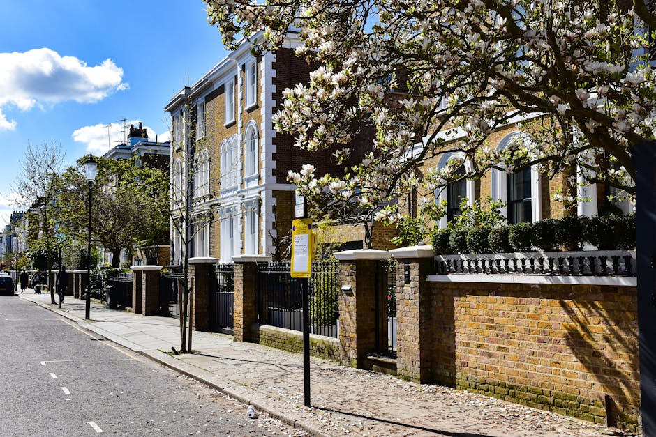 The image depicts a quiet residential street in Hammersmith W6 during daylight, featuring a row of multi-storey Victorian-style buildings with cream-colored facades, decorative molding, and wrought iron balconies. Several large cardboard boxes, some wrapped in plastic, are stacked on the sidewalk near the entrances, indicating packing for a home relocation. A wooden furniture piece and several smaller packed items are visible among the boxes. In the background, a white vehicle is parked, and further down the street, a black van with open rear doors is positioned near a loading area on the pavement, suggesting furniture transport and loading activities. Nearby, a few pedestrians walk along the sidewalk, and mature trees provide shade, contrasting the urban environment. This scene captures an active moving and packing process, consistent with professional removals services like those provided by Hammersmith Man and Van, specializing in home relocation and furniture transport, set against a typical London street environment with clear weather and natural lighting.