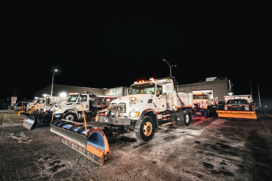 A row of large snowplow trucks equipped with wide metal snow blades attached to the front, parked in an outdoor lot at night under bright streetlights. The trucks are white with orange and black accents, and are positioned side by side on a wet, dark asphalt surface with visible patches of snow and ice. The scene captures the trucks in a stationary state, possibly ready for clearing operations, with some trucks featuring additional equipment such as salt spreaders mounted on the rear. Surrounding the vehicles are commercial buildings and streetlights, illuminating the area. For a home relocation or furniture transport service, such vehicles are typically used for transport or clearing access routes, though here they are seen in a preparation or storage setting relevant to relocation logistics, as carried out by companies like Hammersmith Man and Van.