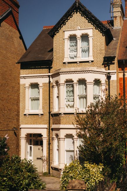 A three-storey Victorian-style house with a light yellow brick facade and white decorative window surrounds, located on a narrow street with neighboring brick buildings. The house features four double-hung sash windows with lace curtains, three on the second floor and one on the third floor, and a front door with a small window and white framing. A paved pathway leads to the entrance, bordered by green shrubs and small trees. The roof is pitched with dark tiles, and a chimney is visible. The scene is illuminated by bright sunlight, with shadows cast on the building, indicating a clear day. The image illustrates the type of residential property often involved in house removals or relocation services, highlighting the exterior environment that [COMPANY_NAME] might service for home moving and furniture transport projects in the Hammersmith area.