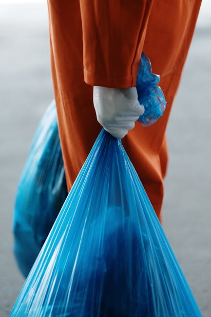 Close-up of a person holding a blue plastic trash bag with their right hand, which is covered by a white glove, during a home relocation or moving process. The individual is wearing an orange jumpsuit, and the background is blurred, suggesting an outdoor setting or loading area. The bag appears to contain waste or packing materials, possibly related to cleaning or clearing space during a house move. This scene may be part of the packing and loading activities facilitated by Hammersmith Man and Van, a removal service specializing in house relocations and furniture transport. The image emphasizes the disposal or temporary storage aspect of moving logistics, with focus on the handling of waste or packing materials, and the action of carrying the bag towards a vehicle or storage area as part of the moving process.