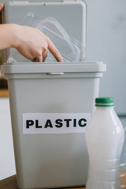 A person placing a clear plastic container into a white recycling bin labeled 'PLASTIC'. The bin is situated on a flat surface indoors, with a second plastic bottle positioned nearby. The scene depicts a home environment focused on waste disposal and recycling, which is an essential aspect of house removals and packing processes. This image highlights the sorting of recyclable materials, supporting eco-friendly relocation practices as part of a comprehensive moving service offered by Hammersmith Man and Van. The lighting is bright and even, emphasizing the cleanliness and organization of the space, with a clear focus on packing materials being sorted for disposal or recycling during the move.